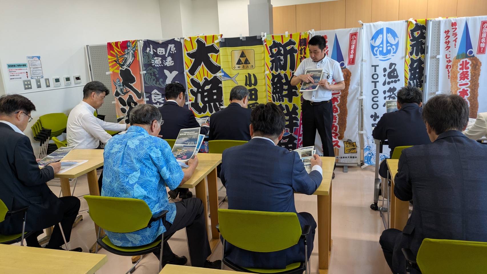 前方の掲示ボードに沢山の商店ののぼり旗がかけられた室内で、複数の参加者がテーブルについている前で、白いシャツを着た一人の男性が手に持った冊子を広げて説明している視察の様子の写真