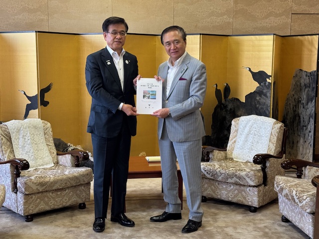 2客のソファが斜めに向かい合って設置された室内に、岩にとまったり飛んでいる黒い鳥が描かれた金屏風を背景に、スーツ姿の市長と知事が中央に立ち一緒に要望書を持っている写真