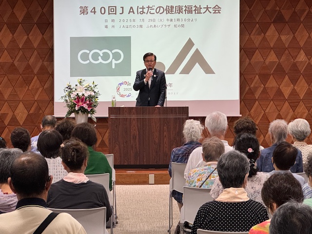 「第40回JAはだの健康福祉大会」と表示されたスクリーンを背景に、ホールに設置された演台に立つスーツ姿の市長が、多くの聴衆を前にマイクを手に挨拶している様子の写真