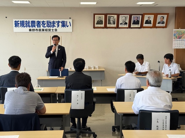「新規就農者を励ます集い」と書かれた横断幕が掲げられている会議室で、関係者や参加者が席に座って見ているなか、前方に立つスーツ姿の市長がマイクを使って挨拶を行っている写真