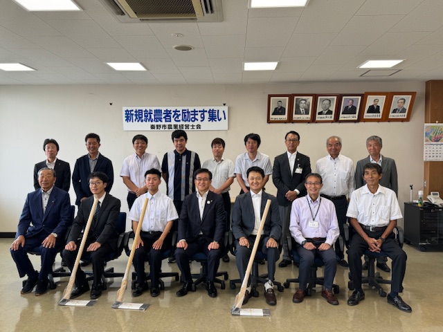 「新規就農者を励ます集い」と書かれた横断幕が掲げられた白い壁の前で、スーツ姿や普段着の男性らが二列に並び、手列の3人の前に真新しい鍬が置かれている記念写真