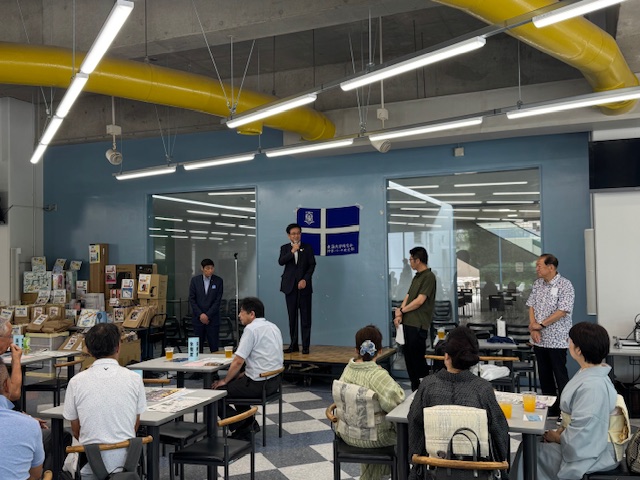 地元の特産品と思われる陳列棚が見え、青い旗が貼られた水色の壁の広間に集まった多くの聴衆を前に、壇上のスーツ姿の市長がマイクで挨拶をしている写真