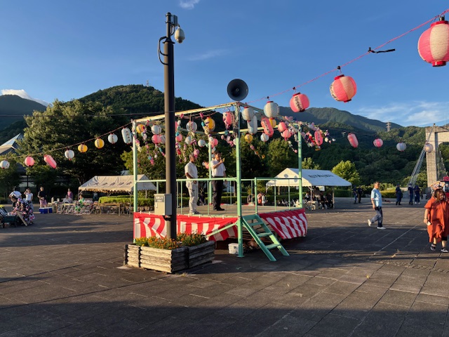太陽が沈みかけた山々に囲まれた祭り会場に、白いテントやカラフルな色の提灯が飾られ、紅白幕がかけられた盆踊りの櫓の上に男性2人が立ち、マイクを持った市長が挨拶をしている写真