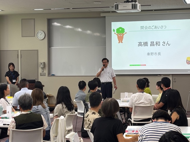 教室前方に「開会のごあいさつ」と「高橋 昌和さん 秦野市長」と表示されたプロジェクタースクリーンが設置され、丸テーブルの席に座った多くの若者と大人を前に、スクリーンの前に立つ白いシャツ姿の市長がマイクを手に挨拶を行っている様子の写真
