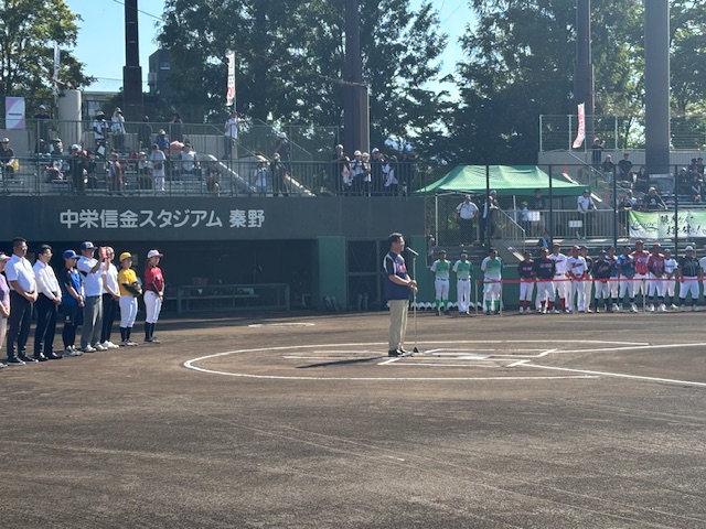 夏の太陽が照りつける野球場で、白いラインが引かれたグラウンドに立つ人々や観客席から見守っている人々のなか、紺色のシャツを着てバッターボックスに立つ市長が開会式の挨拶を行っている様子の写真