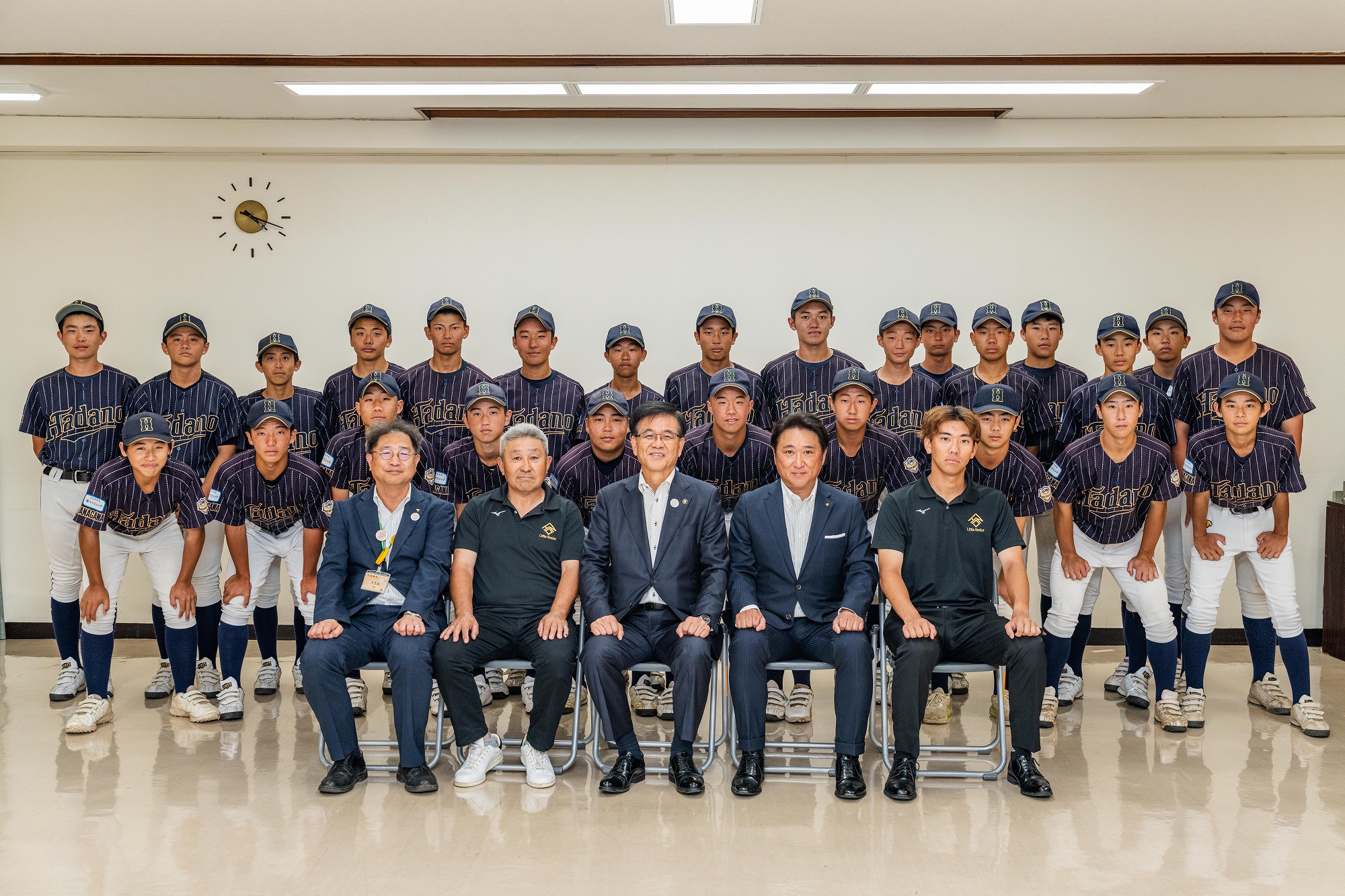時計が設置された白い壁の室内で、市長を含めたスーツ姿の3人の男性と黒いポロシャツを着た2人の関係者が前列の椅子に座り、ストライプ柄の少年野球チームのユニフォームを着た少年たちが2列に並び、笑顔でカメラにポーズをとって集合写真を撮っている様子を捉えた写真
