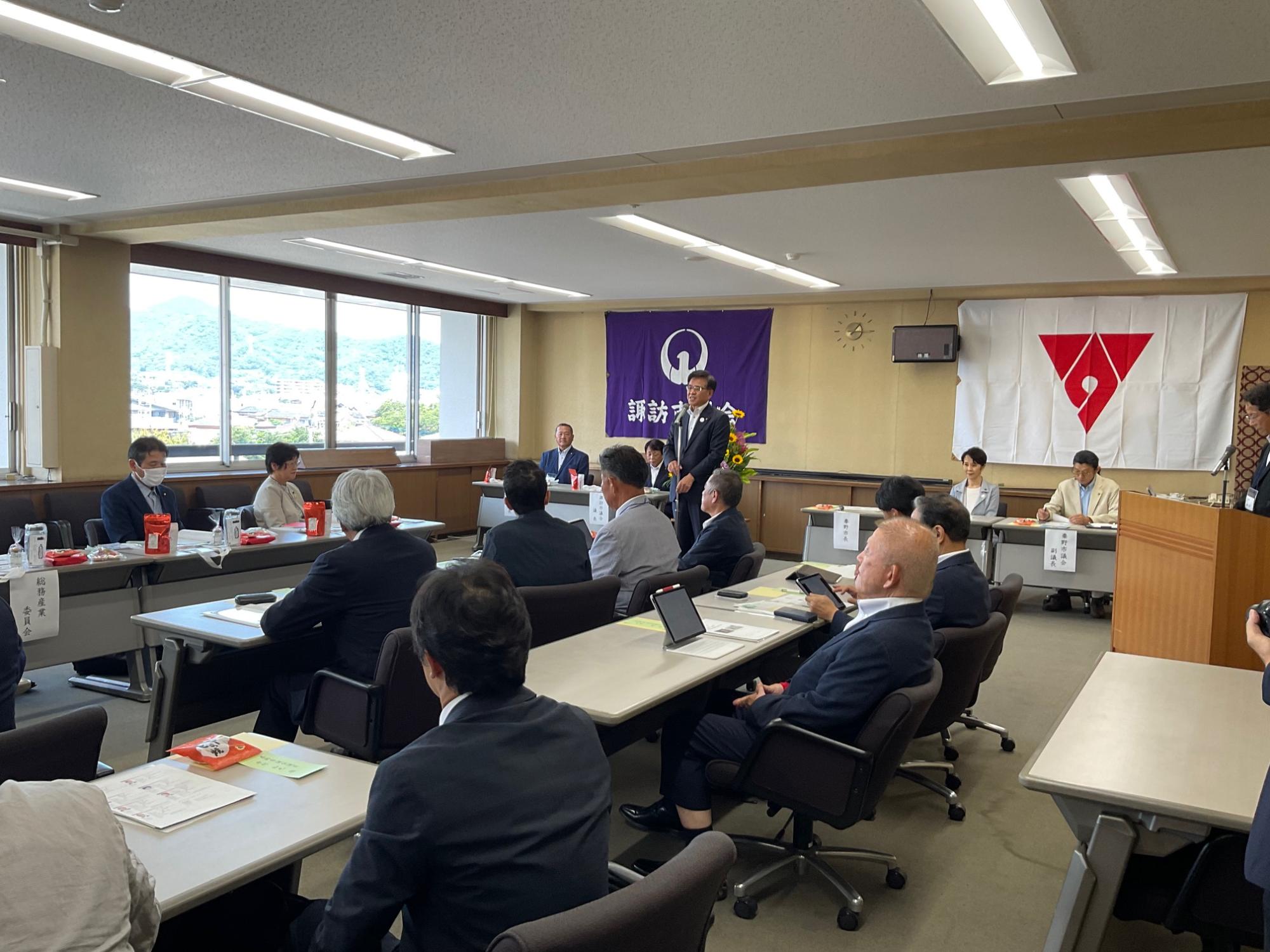 2枚の大きな旗が掲げられた大会議室で、スーツ姿の市長が挨拶をしており、複数のスーツ姿の参加者が机に座って話を聞いている様子の写真
