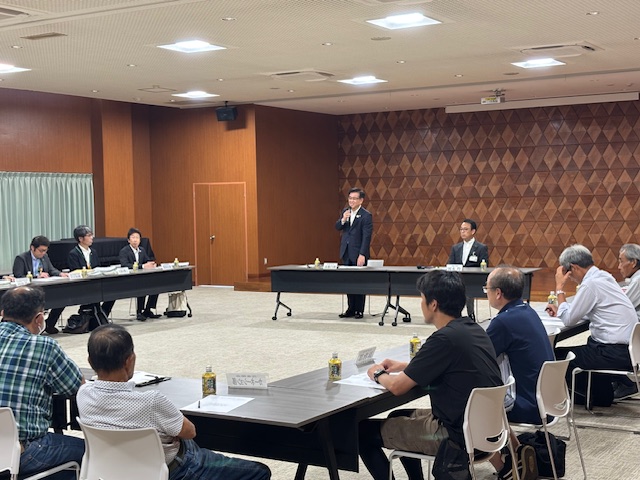 コの字型に並べられたテーブルが設置された大会議室で、中央前方に立つスーツ姿の市長がマイクを持って挨拶を行っており、席に座った多くの参加者が話を聞いている会議の様子の写真