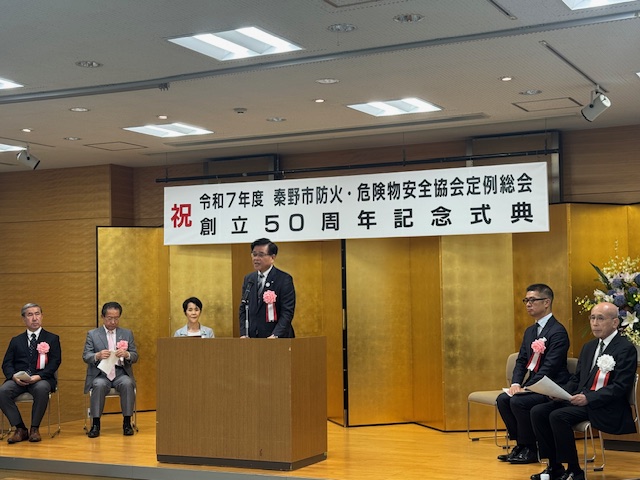 創立50周年記念式典で「祝」と書かれた横断幕の下、左胸に胸章リボンをつけた来賓、金屏風や花飾りが写っている檀上の中央で、演台に立つスーツ姿の市長が挨拶を行っている写真