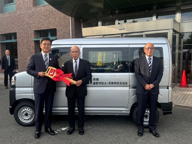 消防安全協会が消防本部に寄贈したグレー色の軽バン型の広報車を前に、スーツ姿の市長と2人の関係者の男性が赤い巨大な鍵を手に記念撮影をしている様子を写した写真