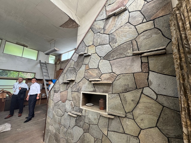 2階へ上がる階段の壁が不揃いの形をした石が積み重なって作られた室内を、白いシャツに黒いパンツ姿の市長と2名の男性が視察している様子の写真
