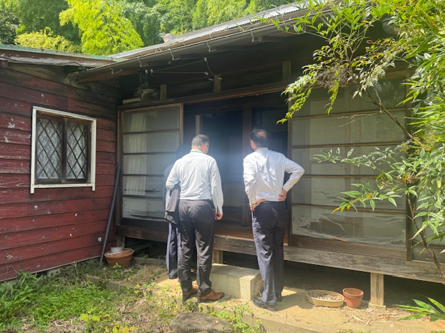木造の壁や格子窓、傷んだ雨戸が特徴的な古い日本家屋の中を、白いシャツ姿の二人の男性が庭先から立って見ている写真