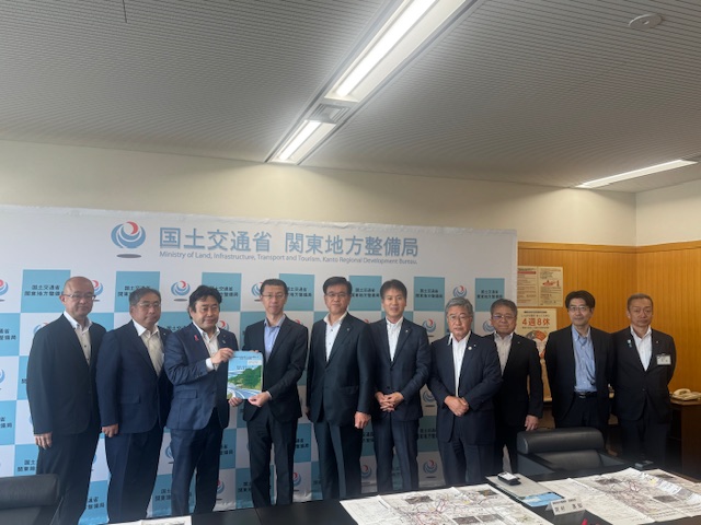 国土交通省関東地方整備局のロゴが描かれたパネルを背景に、10人のスーツ姿の男性が横一列に並び、左から3番目と4番目に立つ2人の男性が要望書を一緒に持ち笑顔で集合写真を撮っている写真