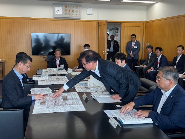 黒い横長のテーブルを囲むスーツ姿の男性たちが広げられた地図を囲み、スーツ姿の市長が地図上の一点を指差しながら真剣に説明をしている様子を捉えた写真