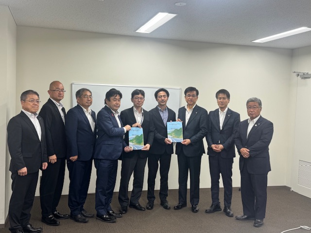 ホワイトボードが設置された白い壁の部屋で、スーツ姿の9人の男性が横一列に並び、中央に立つ4人の男性が2人ずつ一緒に資料を手に持ち立っている集合写真