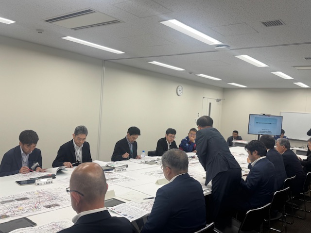 多くの男性が大きなテーブルを囲んで座っており、テーブルに広げられた大きな地図や資料を前にして、手前中央の席から立ち上がったスーツ姿の男性が説明をしている様子を捉えた会議の様子の写真