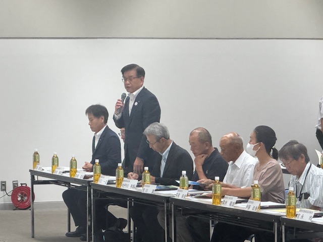 席札やペットボトルのお茶が置かれた机と椅子が並んだ会議室で、スーツ姿の市長がマイクを持って立ち、横に座る出席者たちを前にスピーチを行っている様子の写真