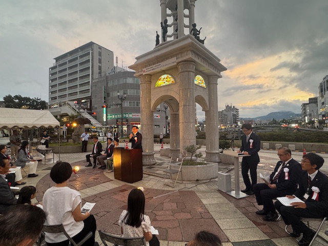 夕焼けの空が広がる屋外広場で、半円形の窓が黄色やオレンジ色にライトアップされた石造りのモニュメントの前に設置された檀上に立つスーツ姿の市長の挨拶を、椅子に座っている周囲の人々が話を聞いている点灯式の様子の写真