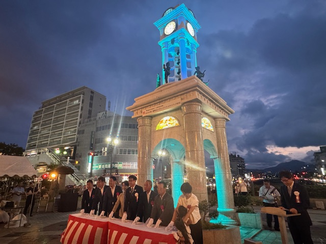 太陽が沈んだ夜空の下、青色にライトアップされた時計台の前で、複数のスーツ姿の関係者が紅白幕で囲われた台に手を置き、点灯式を行っている様子を撮影した写真