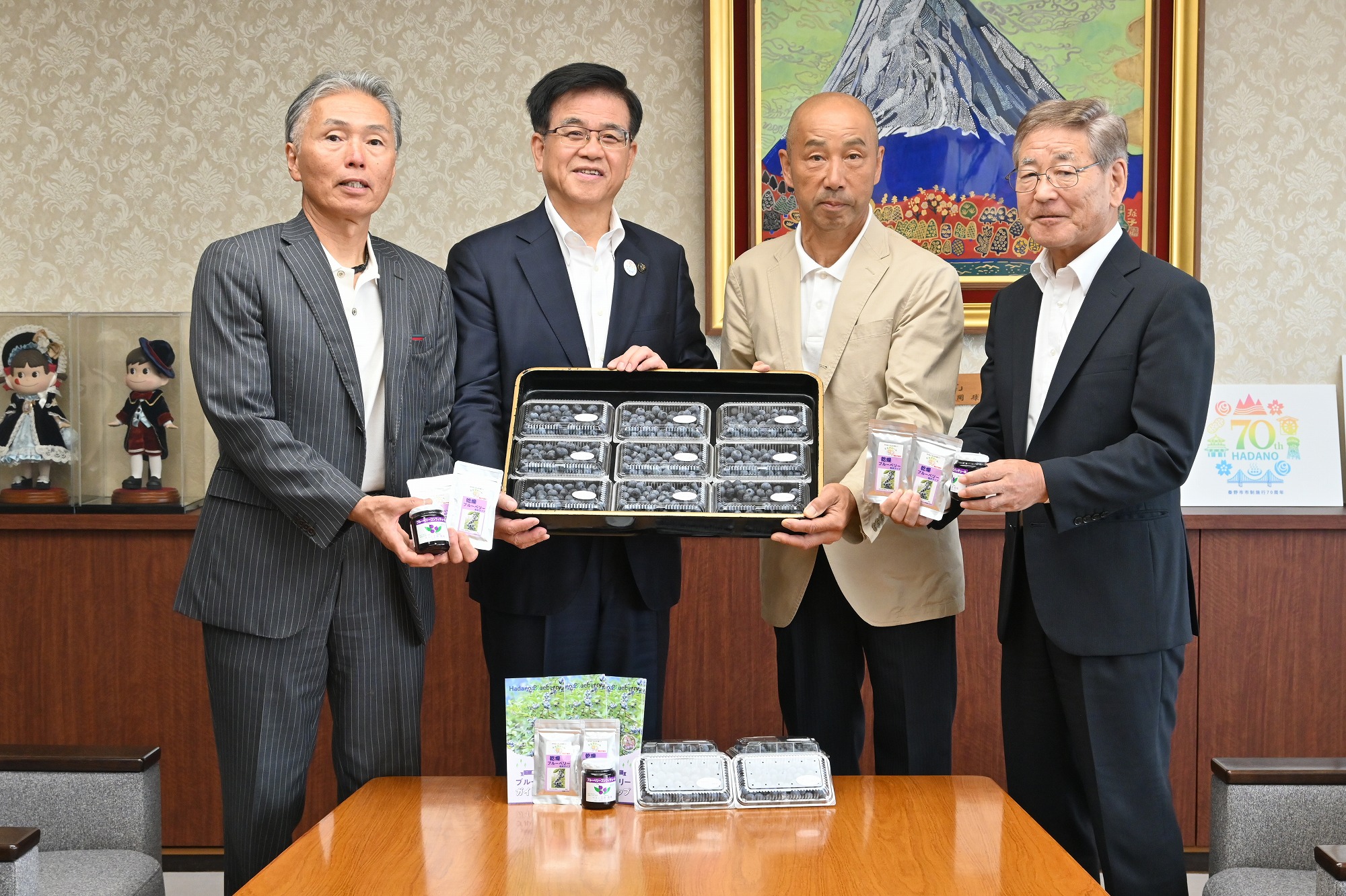 金色の額縁に入った山頂に雪が積もった大きな山の絵画が壁に飾られた落ち着いた背景の部屋で、様々なブルーベリーの商品が置かれた木製のテーブルを囲んでスーツ姿の市長を含め4人の男性が、黒いお盆に入った9つの透明パック入りのブルーベリーを手に持ち記念撮影をしている様子を写した写真