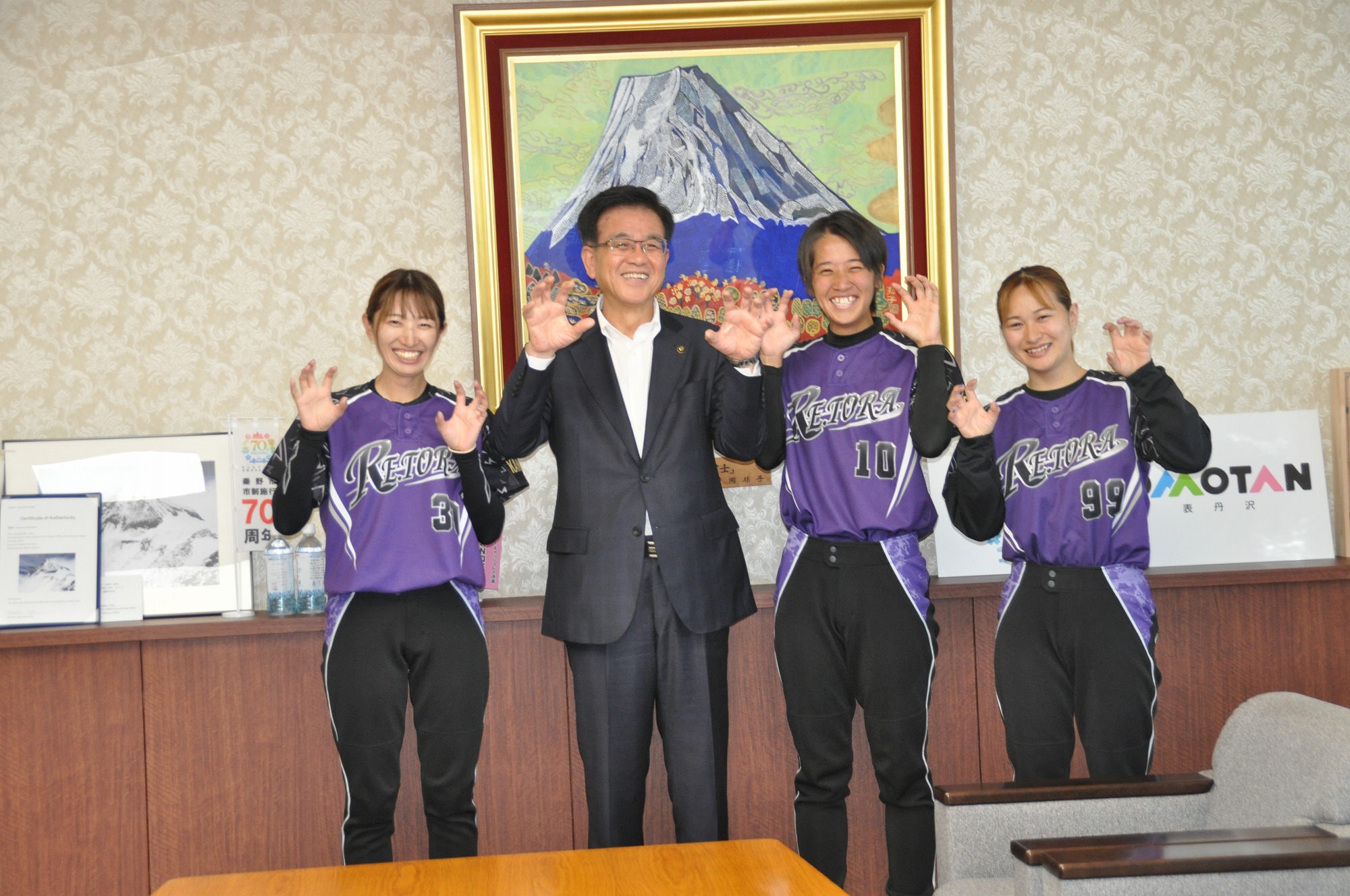金色の額縁に入った山頂に雪が積もった大きな山の絵画が壁に飾られた落ち着いた背景の部屋で、スーツ姿の市長が紫色のユニフォームを着た三人の女性選手と並んで笑顔で両手を上げている記念写真