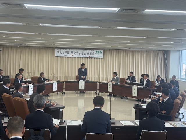 「令和7年度県央やまなみ協議会」と書かれた横断幕が掲げられた会議室で、中央に立つスーツ姿の市長が挨拶をしており、円形に設置された席に座った関係者が真剣な表情で話を聞いている会議風景を写した写真