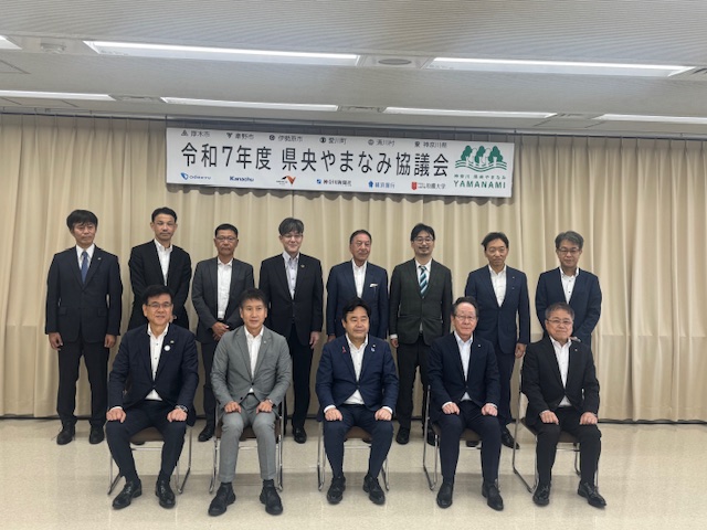 白い緞帳を背景に「令和7年度県央やまなみ協議会」と書かれた横断幕の下で、多くのスーツ姿の男性が2列に並んで、集合写真を撮影している様子の写真