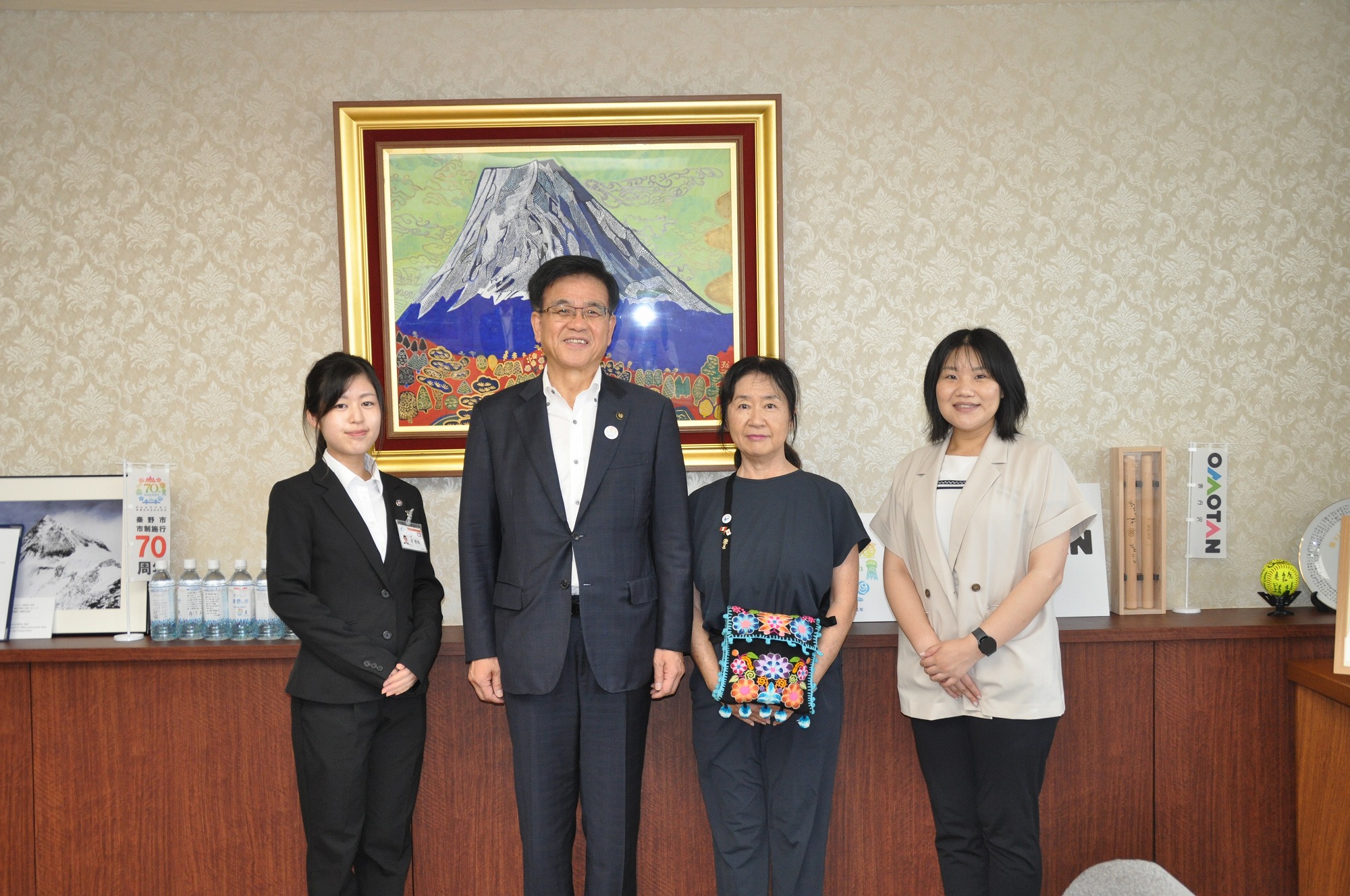 大きな山の絵画が壁に飾られた落ち着いた背景の部屋で、スーツ姿の市長を囲み、ビジネススーツとカジュアルな服装の3人の女性たちが記念撮影に応じる様子を捉えた写真