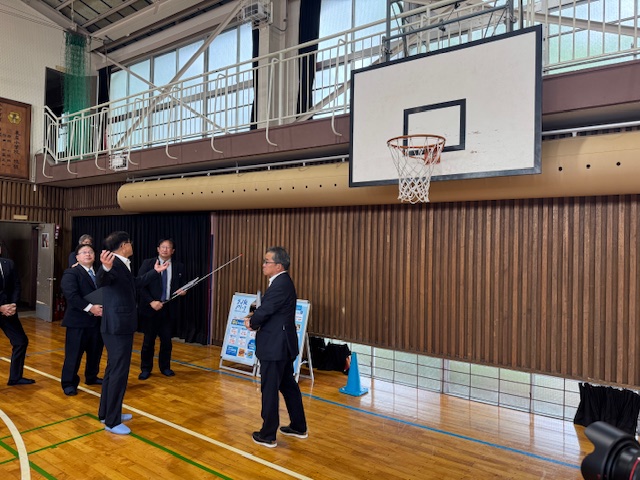 背景にはバスケットボールのゴールや木製の壁のある体育館で、集まったスーツ姿の男性たちに、市長が説明をしている様子の写真