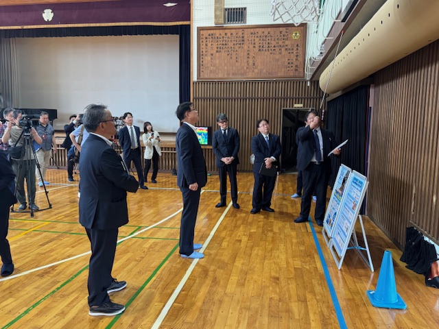 体育館の天井から吊り下げられた空調設備を男性が指差し、大勢の男性たちが見学しいる様子を複数のカメラマンが撮影している視察風景の写真