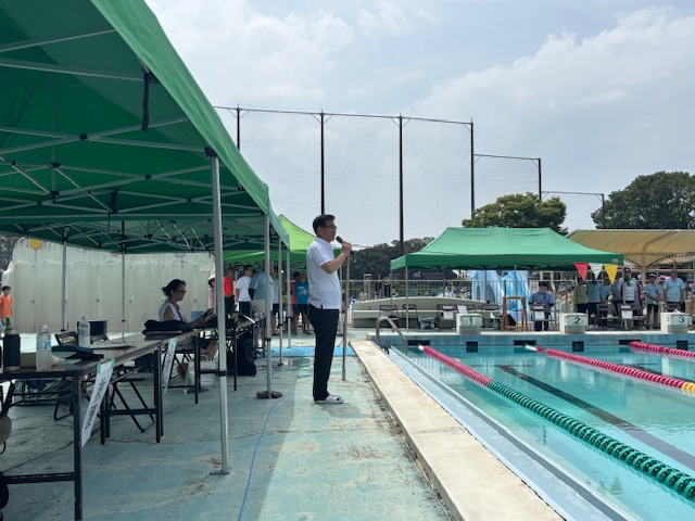 緑色のテントが並ぶ屋外プールサイドに立つ白いシャツ姿の市長がマイクを持ち挨拶をしており、多くの参加者が見守っている水泳競技大会の開会式の様子を捉えた写真