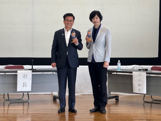 舞台上で、スーツ姿の市長と合田雅吏さんが長机の前に立ち、それぞれが二宮金次郎を模した人形を持ってカメラに向かい、笑顔で記念撮影をしている写真