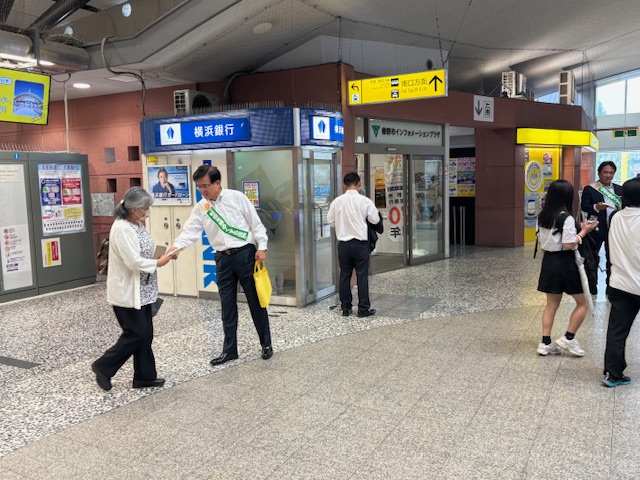 駅構内でたすきをかけた市長が、年配の女性に笑顔でチラシのようなものを手渡し、周囲には他のスタッフや利用者が行き交う風景を写した啓発活動の様子の写真