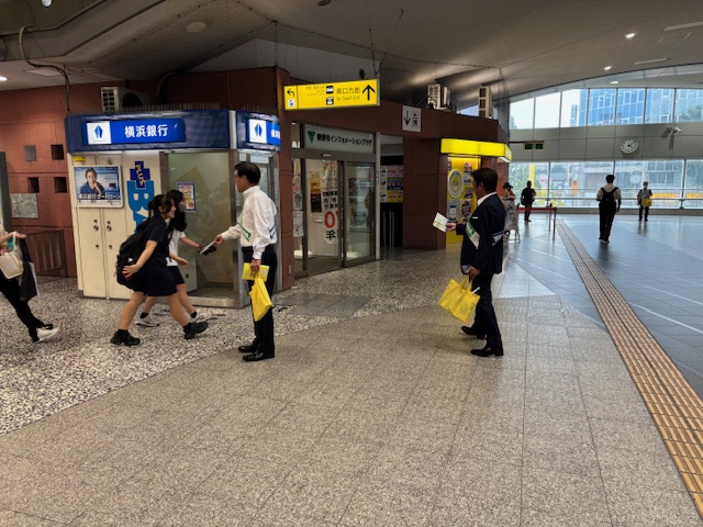駅構内で、スーツ姿の男性や白いシャツを着た市長が黄色い配布物を持って、女子高生や通行人に声をかけており、多くの人々が忙しそうに行き交う様子を捉えた啓発活動の様子の写真