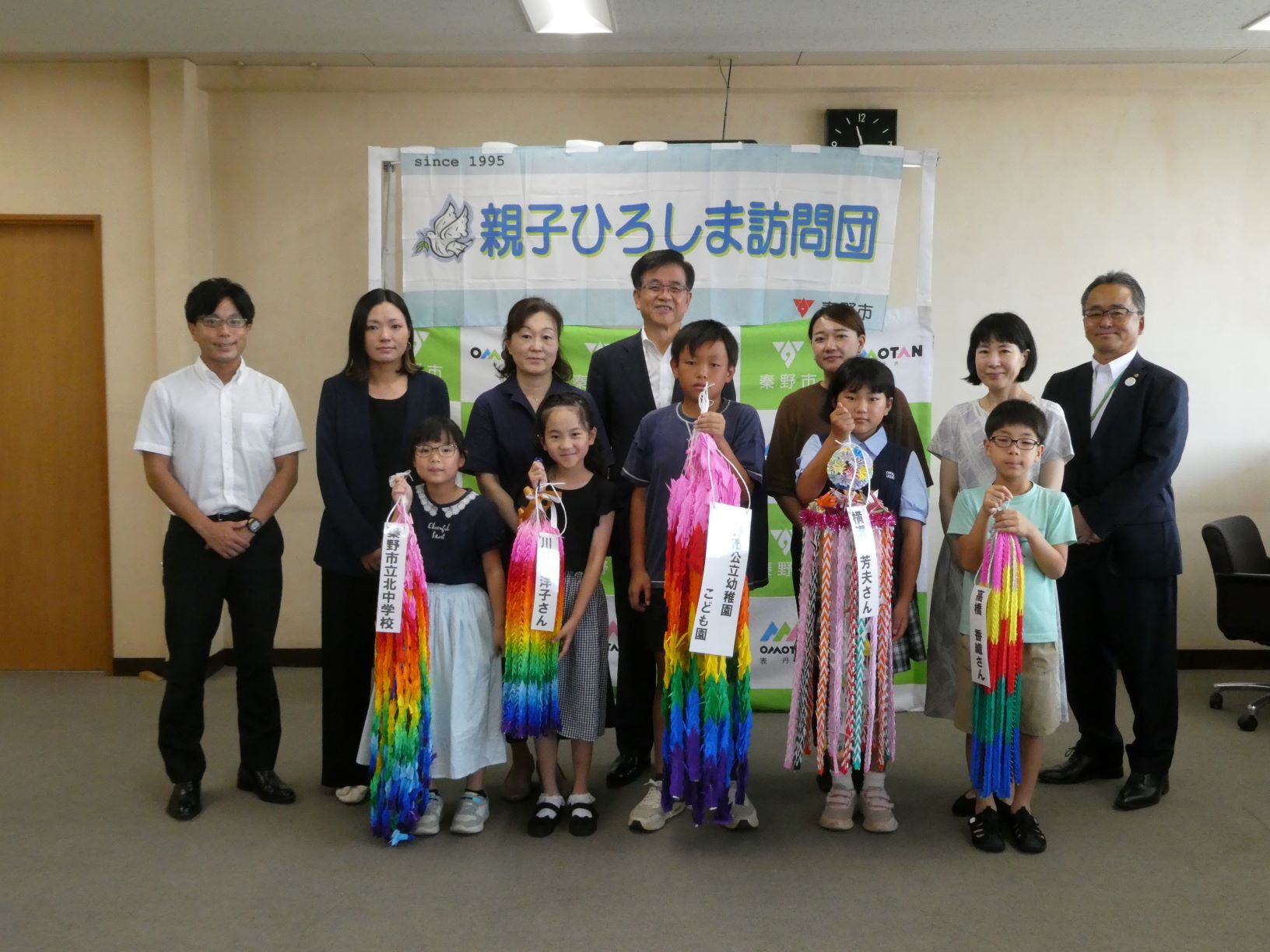 「親子ひろしま訪問団」と書かれた横断幕とバックパネルを背景に、5人の子供と市長や大人が並び、色とりどりの千羽鶴を手に笑顔で記念撮影をしている様子の写真