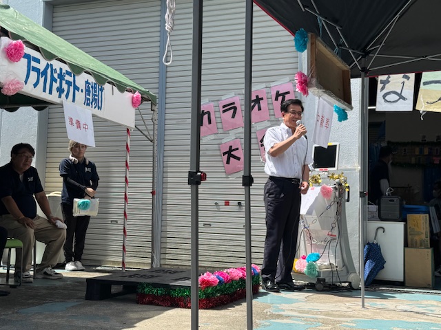 カラオケ大会と書かれた看板やピンク色の飾り付けがされた屋外イベント会場のテントの下で、白いシャツ姿の市長がマイクを片手に挨拶を行っている様子の写真