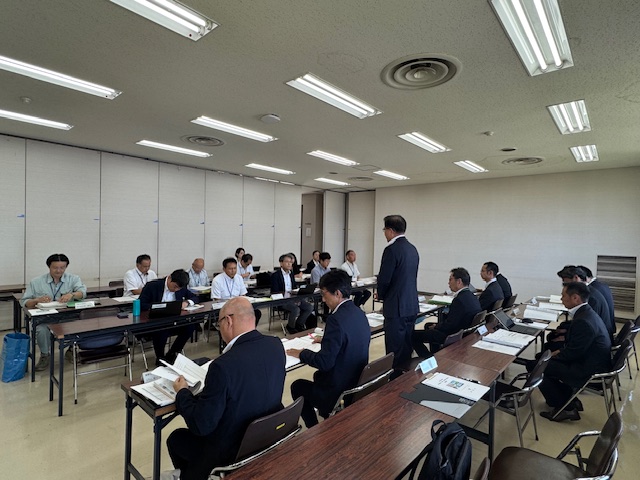 大会議室で多くのスーツ姿の男性が机に座って資料に目を通す中、中央に立つスーツ姿の市長が県の関係者に向けて説明をしている様子の写真