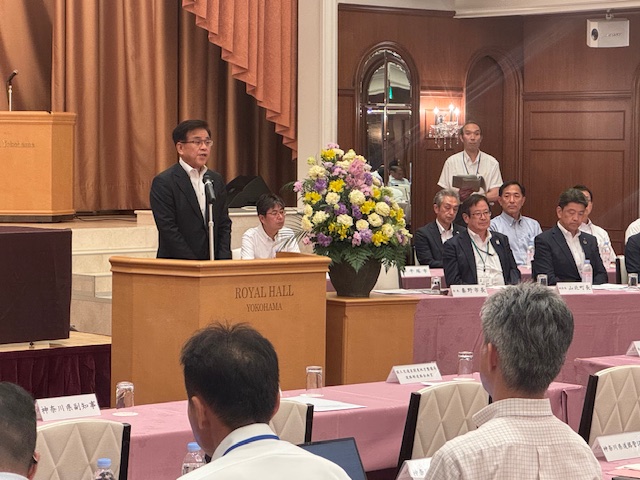 華やかな装花が飾られた会場で、スーツ姿の市長が木製の演台に立ち、多くの来場者に向けて挨拶をしている様子の写真