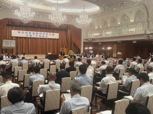 「神奈川県道路利用者会議」の横断幕が掲げられた大きなホールで、演壇に立つスーツ姿の市長が、多数の参加者に向けて挨拶をしている会議の様子を捉えた写真