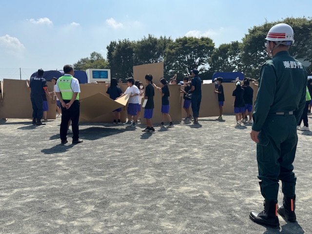 広大なグラウンドで、数名の引率者と複数の児童・生徒がダンボールを用いて壁やブースのような構造物を共同で組み立てている防災訓練の様子の写真