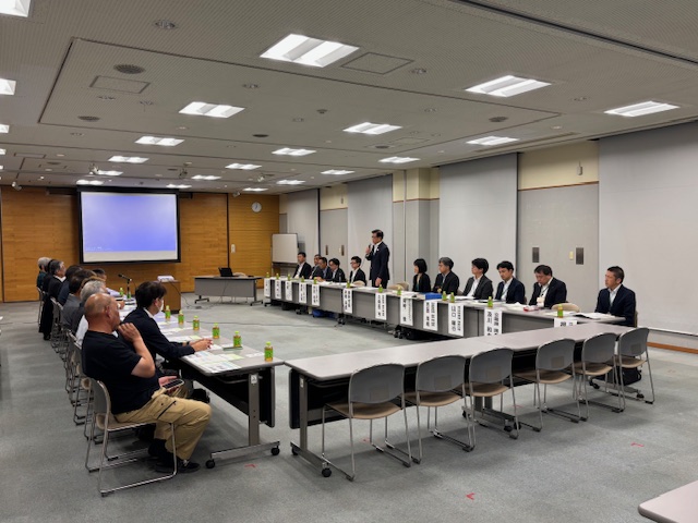 広い会議室で、スーツ姿の男性がコの字型に並べられた机の前に立ち、多くの出席者に向かって熱心に発言しており、奥にはプロジェクターのスクリーンが見える懇親会の様子の写真