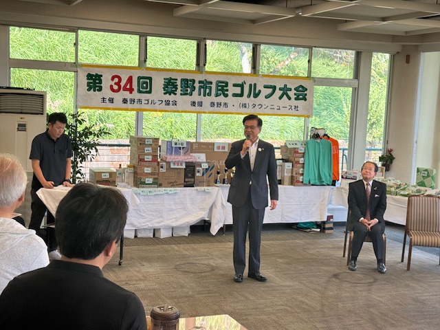「第34回 秦野市民ゴルフ大会」と書かれた横断幕の下、景品が山積みにされたテーブルの前で、スーツ姿の市長がマイクを持ち、着席した参加者に向けてスピーチをしている表彰式の様子の写真