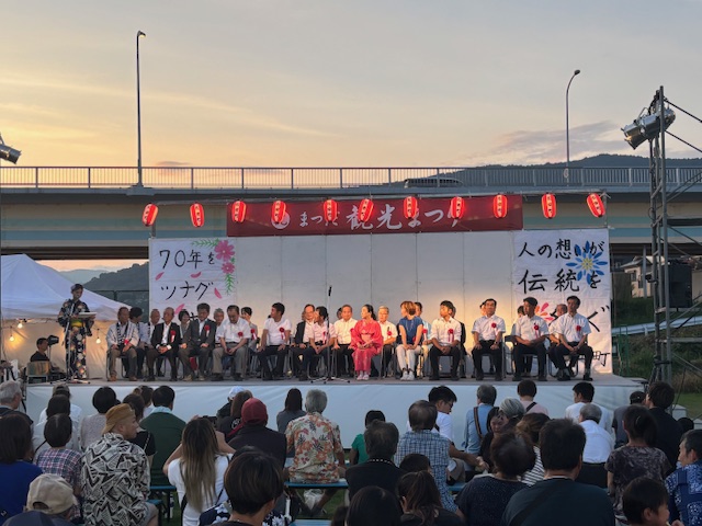 夕焼け空の下、川沿いと思われる橋の下の特設ステージで、浴衣姿の司会者がマイクを持ち、大勢の観客を前に、多くの来賓らしき人々が横一列に並んで着席している祭りの様子の写真