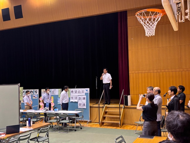 体育館のステージ上で、白いシャツ姿の市長がマイクを持ち、数名の聴衆や関係者が囲む中でスピーチをしており、ステージ下には数名の男性と「希望の丘」「生活訓練」などのパネルが見える様子の写真