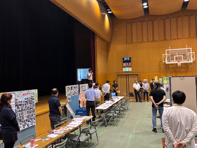 体育館のステージ上で、白いシャツ姿の市長がマイクを持ち、床にはテーブルと椅子が並べられ、複数の関係者が各ブースの前に立ち話を聞いている写真