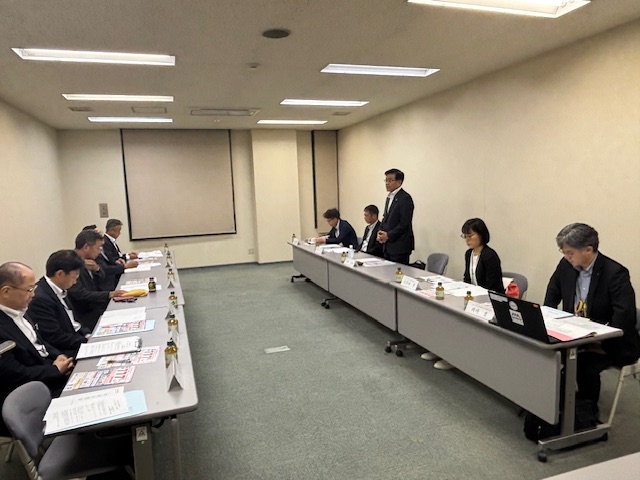 会議室の長机が向かい合わせに配置され、中央のスーツ姿の市長が起立してスピーチをしており、多数の出席者が着席してそれに耳を傾けている会議の様子の写真