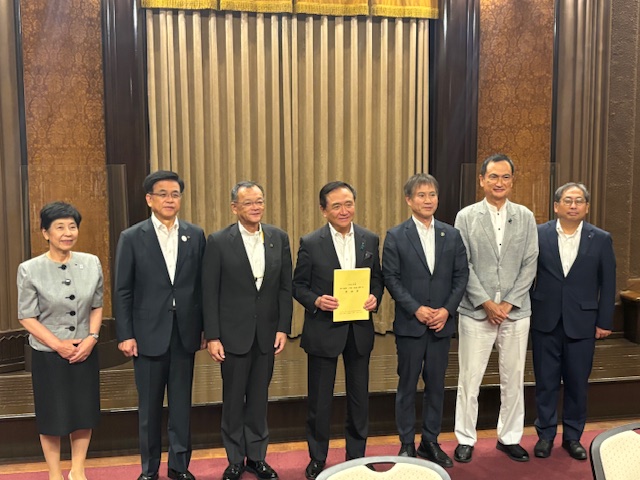 カーテンが飾られた会場のステージ上で、スーツ姿の女性1名と男性6名が並んで立っており、中央の男性が黄色の冊子のようなものを手に持っている集合写真