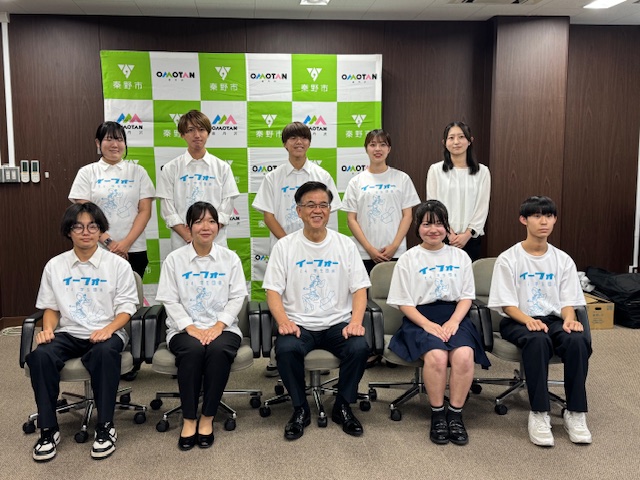 「秦野市」のバックパネルある部屋で、中央の白いTシャツ姿の市長を囲み、同じTシャツを着た4名の男性と5名の女性が立ったり座ったりして並んでいる集合写真