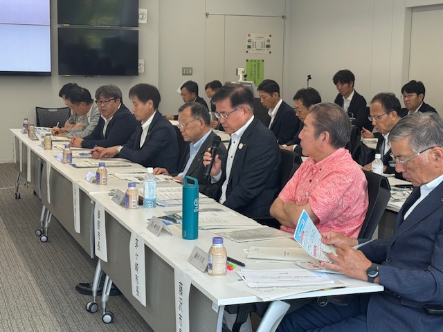 長机を囲んで座る多くの出席者がいる会議室で、中央のスーツ姿の市長がマイクを持ち、熱心に発言しており、他の出席者は資料を確認している様子の写真