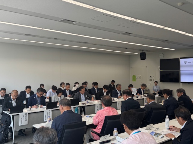 会議室でコの字型に配置された机を囲み、多数のスーツ姿の男性を中心とする出席者が、資料を確認したりメモを取ったりしている懇談会の様子の写真